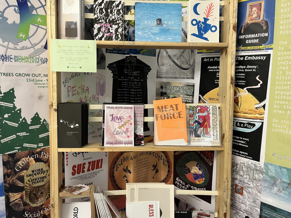 A collection of colourful pamphlets sits on an open wodden shelf in front of a wall covered in colourful art show posters